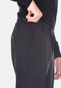 Uomo che regola la vita di pantaloni neri con cerniera e bottone, indossando un top nero a maniche lunghe, su uno sfondo chiaro.