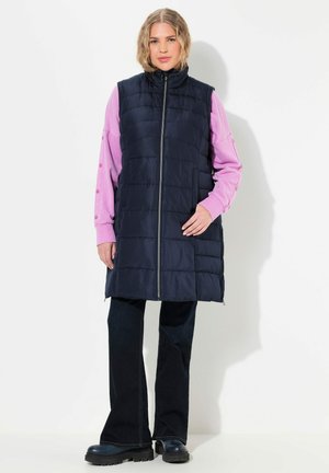 Chalecos acolchados en navy con cuello alto y cremallera frontal. Combinado con una blusa rosa de manga larga que cuenta con detalles de botones. Jeans oscuros acampanados y botas gruesas.