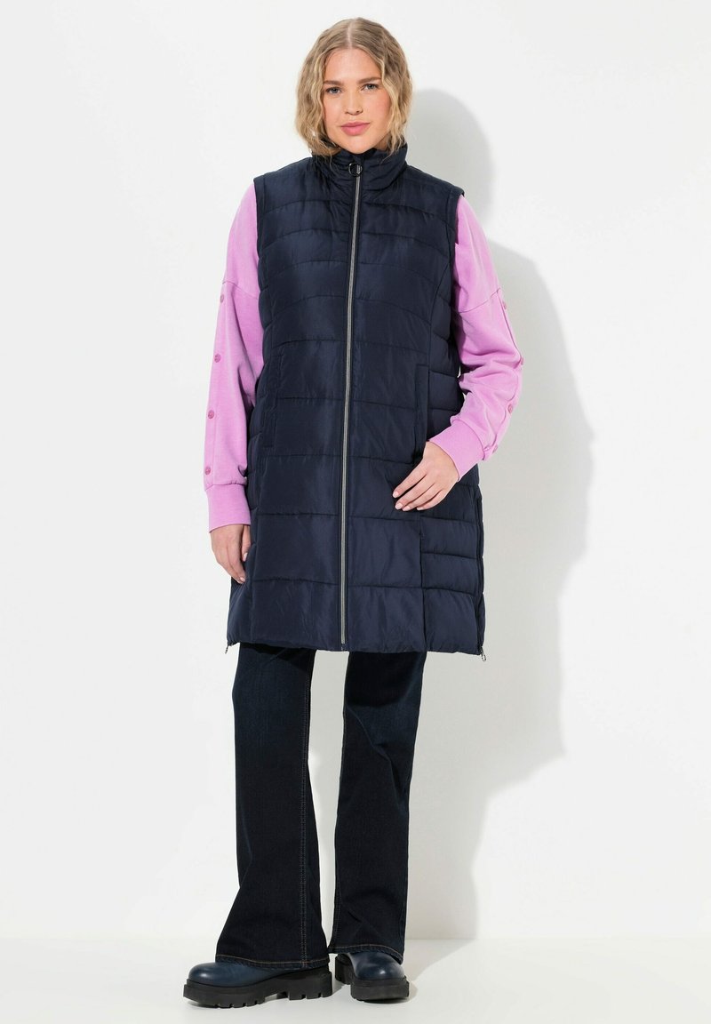 Marinblå vadderad väst med hög krage och frontdragkedja. Kombinerad med en rosa långärmad topp med knapprad. Mörka utsvängda jeans och kraftiga stövlar.