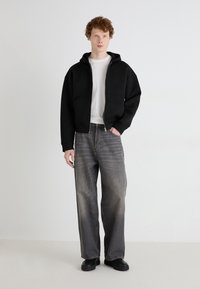 Haut de survêtement zippé noir avec une capuche et des poches avant, associé à un jean gris clair, décontracté et ample. Le modèle se tient dans un décor neutre.