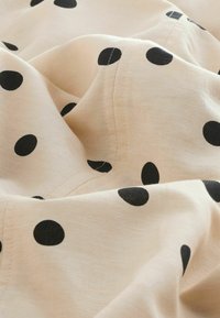 Vaaleanbeige kangas, jossa on mustia pilkkuja, joka omaa sileän tekstuurin ja pehmeän laskostuksen, näyttäen saumat ja taitteet koko materiaalissa.