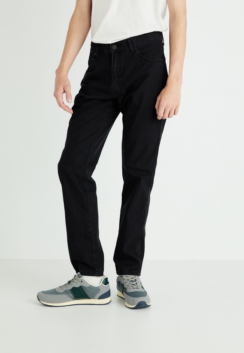 Jeans negre din denim cu un croi clasic, având talie cu nasturi, buzunare laterale și o textură netedă. Purtați cu adidași gri și verzi.
