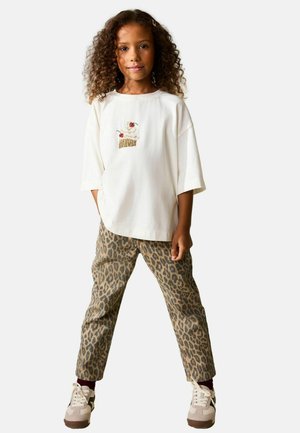 Weißes übergroßes T-Shirt mit einer Cupcake-Stickerei und beige Leo-Hose. Beige Sneaker mit weißen Akzenten. Lockiges Haar.