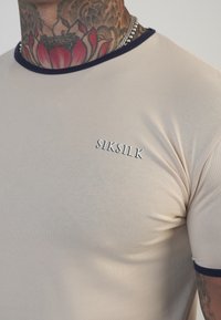 Béžové bavlnené tričko s námorníckym ribbovaným výstrihom, s malým vyšitým logom "SIKSILK" na hrudi, s priliehavým strihom.