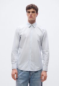 Vaaleansininen pitkähiha-painonappipaita kauluksella, jossa on himmeä kiilto, kuvioitu kangas ja hillitty logo rintakehässä.