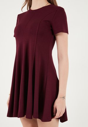 Kobieta w bordowej sukience z krótkim rękawem i rozkloszowanym dołem, z subtelnymi przeszyciami, prezentująca tatuaż na przedramieniu na jednolitym tle.