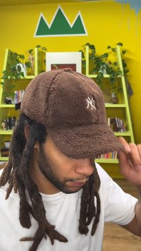 Bruine fleecepet met een geborduurd logo aan de voorkant, een gebogen klep en een verstelbare band aan de achterkant. De achtergrond bevat planten en een gele muur.