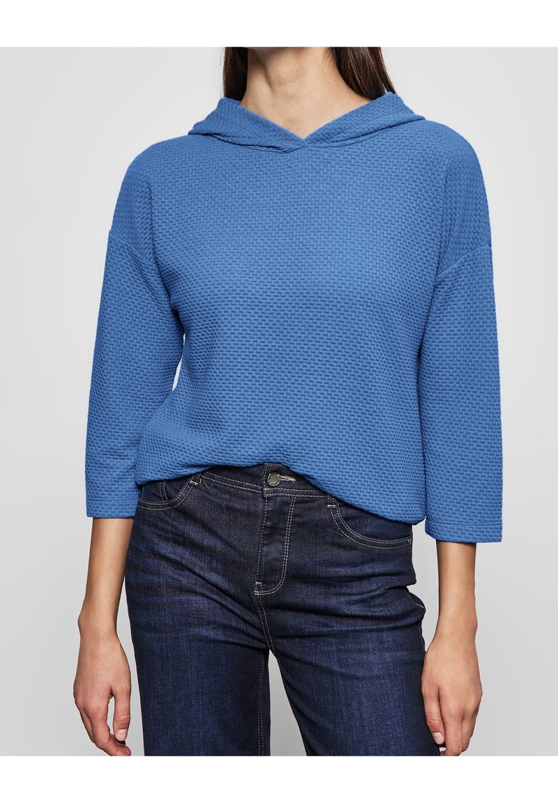 Vrouw die een gestructureerde blauwe hoodie met driekwart mouwen draagt, ingestopt in donkerblauwe denim jeans.