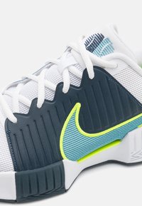 Nærbilde av hvit treningssko med mørkeblå og neongrønne aksenter, som har lisser og teksturert Nike swoosh-logo på mesh-overdelen.