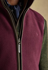 Bordový fleeceový vest s vysokým golierom, kontrastnou tmavozelenou látkou po stranách a kovovým zipsom s označeným ťaháčikom.