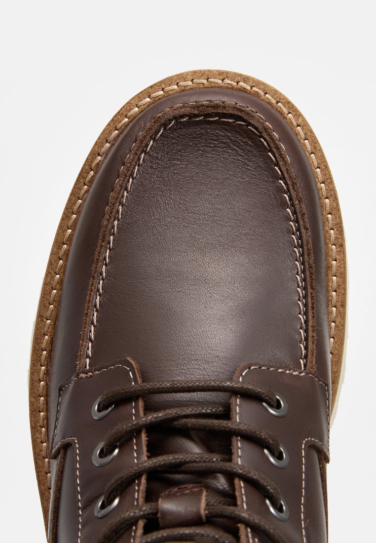 Braune Lederschuhe mit runder Spitze, kontrastierenden Nähten und einer strukturierten braunen Sohle. Die Schnürsenkel sind durch metallene Ösen gefädelt.