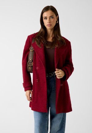 Manteau en laine bordeaux avec un design à double boutonnage, des revers larges et un tissu texturé ; porté sur un haut marron et associé à un jean taille haute.