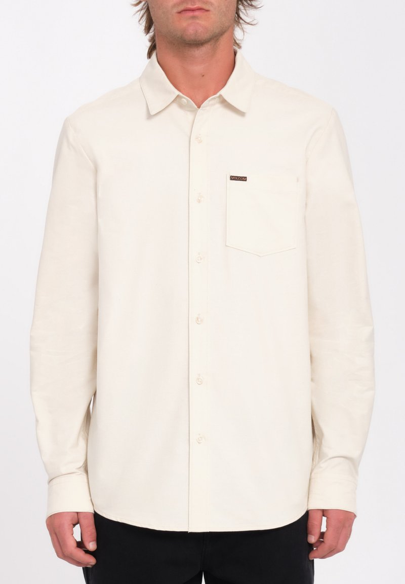 Chemise à manches longues couleur crème avec fermeture à boutons, col et poche poitrine arborant une petite étiquette de marque. Texture de tissu lisse.