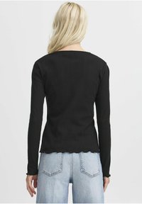 Černý top s dlouhým rukávem, žebrovanou strukturou, vlnkovým lemem a přiléhavým designem. Nesený s světle modrými džíny s vysokým pasem.