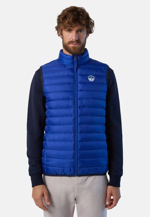 Gilet blu imbottito con cuciture orizzontali, colletto alto e chiusura con zip. Presenta un'etichetta con logo sul lato sinistro. Indossato sopra un maglione scuro.