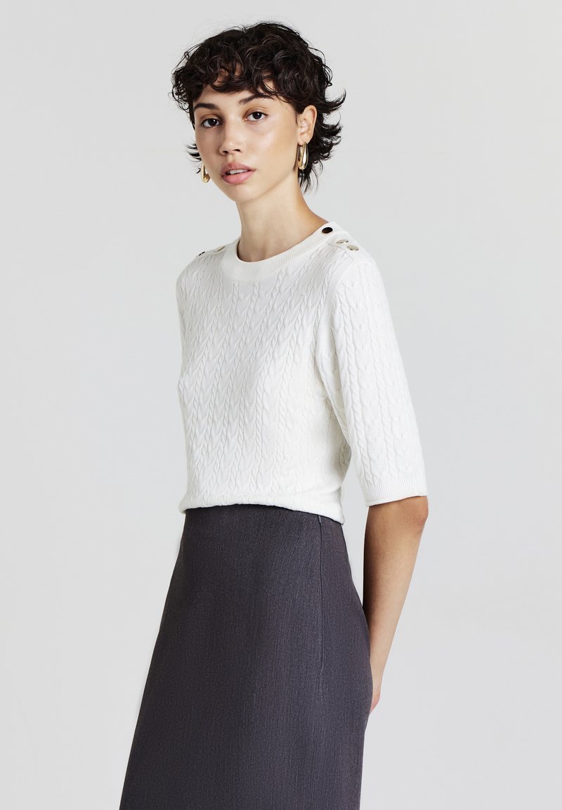 Pull en tricot blanc à manches courtes, avec des boutons noirs sur l'épaule, associé à une jupe gris foncé. Texture lisse.