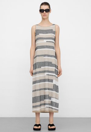 Vrouw draagt een mouwloze, tot op de enkels vallende jurk met een horizontaal gestreept patroon in beige, wit en zwart, zwarte zonnebril en zwarte sandalen met open tenen.