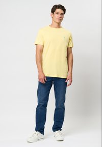 Tricou din bumbac galben, cu guler rotund și mâneci scurte, asortat cu blugi albaștri și adidași albi. Prezintă un mic logo albastru pe piept.
