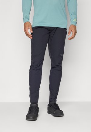 Pantalones deportivos negros con corte ajustado, que presentan un acabado texturizado y acentos de costura. Combinados con zapatos negros y una camisa de manga larga en color azul verdoso.