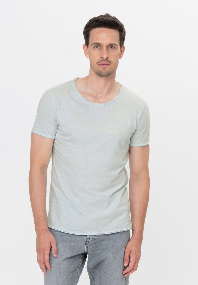 Uomo con capelli corti castani che indossa una maglietta a maniche corte aderente di colore azzurro chiaro e jeans grigi chiari, in piedi contro uno sfondo bianco.