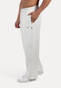 Homme portant un pantalon de survêtement blanc et des baskets blanches, la main reposant dans la poche latérale, se tenant contre un fond clair.
