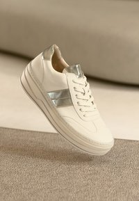 White leather sneaker with silver accents, featuring a round toe, lace-up front, and thick sole. Minimalist design with smooth texture.
