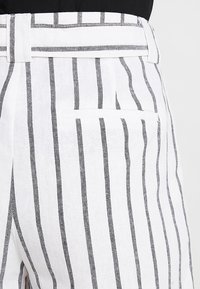 Białe lniane spodnie w pionowe czarne paski, z wyraźnie zdefiniowanym pasem i jedną tylną kieszenią. Materiał wydaje się lekki i teksturowany.