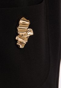 Schwarzer Stoff mit einer strukturierten Oberfläche, auf der sich ein glänzendes, abstraktes goldfarbenes Broschen-Detail an der Seite befindet.