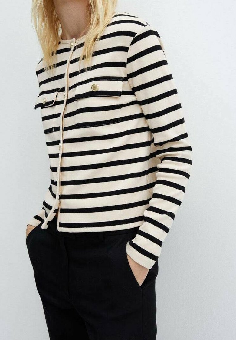 Femme portant une veste à rayures horizontales crème et noire à manches longues avec des poches à boutons dorés, associée à un pantalon noir, mains dans les poches.
