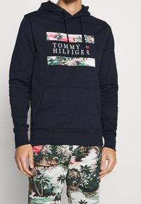 Mężczyzna ubrany w granatową bluzę z kapturem Tommy Hilfiger z tropikalnym wzorem oraz pasujące spodnie z tropikalnym nadrukiem przedstawiającym palmy i chatki.