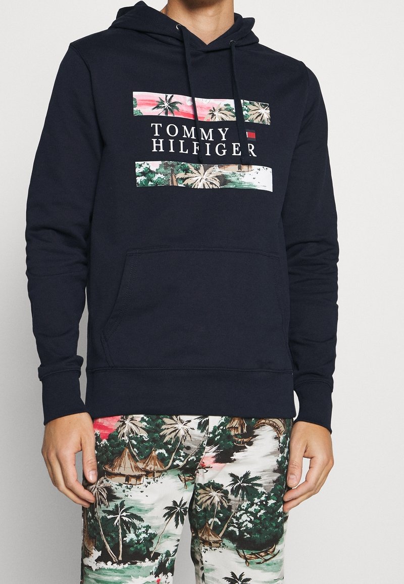 Mężczyzna ubrany w granatową bluzę z kapturem Tommy Hilfiger z tropikalnym wzorem oraz pasujące spodnie z tropikalnym nadrukiem przedstawiającym palmy i chatki.