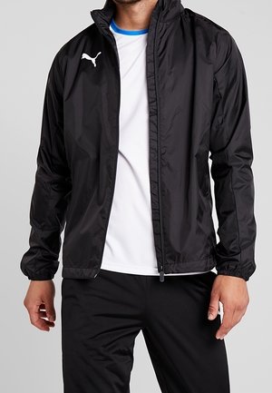 Homme portant une veste zippée noire Puma sur un T-shirt blanc avec un liseré bleu, assortie à un pantalon noir, debout devant un fond uni.