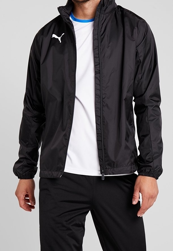 Homme portant une veste zippée noire Puma sur un T-shirt blanc avec un liseré bleu, assortie à un pantalon noir, debout devant un fond uni.