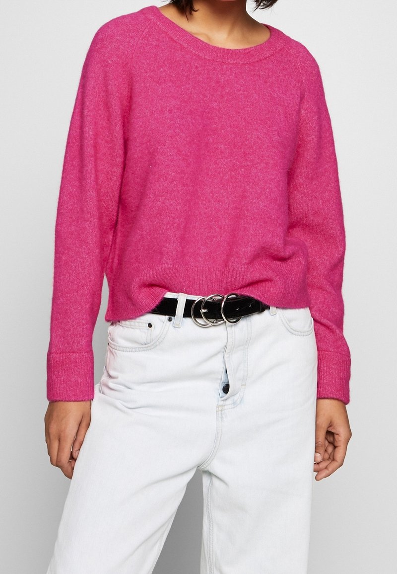 Suéter de punto rosa con mangas largas, escote redondo y puños acanalados, combinado con unos vaqueros de cintura alta de color azul claro y un cinturón negro con herrajes plateados.