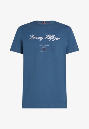 Niebieska koszulka z krótkim rękawem z napisem "Tommy Hilfiger", założona w 1985 roku, Nowy Jork, oraz małe logo na lewym rękawie.