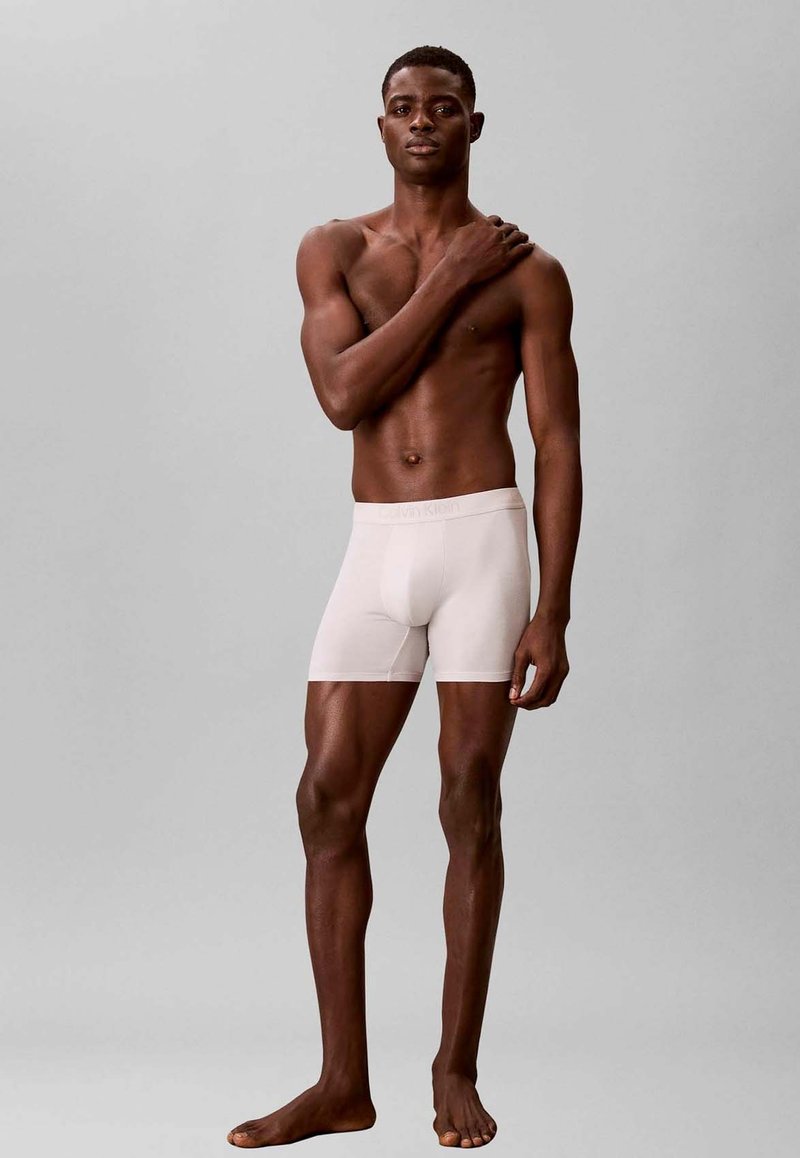 Gespierde man staat blootsvoets op een grijze achtergrond, draagt witte boxershorts, met één hand rustend op zijn tegenovergestelde schouder.