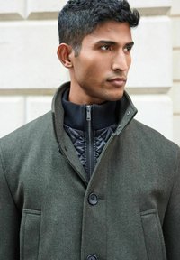 Olivgrüner Fischgrätmantel mit hohem Kragen, ausgestattet mit einem schwarzen Reißverschlussinnenfutter und Frontknöpfen, glatter Textur und seitlichen Taschen.