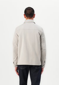 Giubbotto in cotone beige con colletto e maniche lunghe, con bottoni ai polsini, visto dal retro. Jeans scuri visibili sotto.