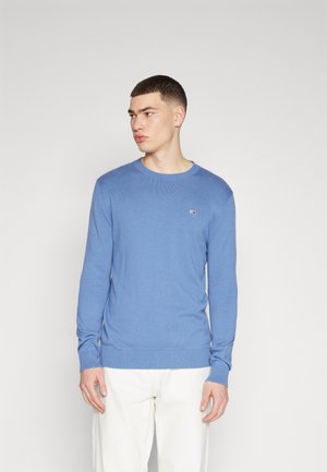 Pull en tricot bleu clair avec des manches longues, un col et des poignets côtelés, arborant un petit accent de logo sur la poitrine. Associé à un pantalon blanc.