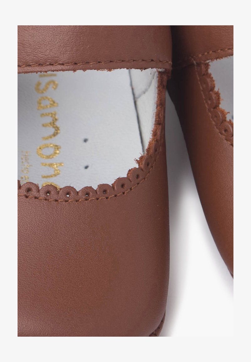 Pantofi din piele maro cu margini ondulate în jurul deschiderilor. Interiorul are căptușeală albă cu textul logo-ului aurit. Textură netedă.