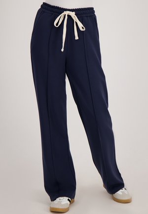Marineblauwe wijde sweatpants met een trekkoord in de taille, gecombineerd met wit en grijze sneakers op een effen achtergrond.