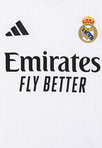 Balts futbola krekls ar melnu Adidas logo, tekstu "Emirates Fly Better" un izšūtu Real Madrid ģerboni ar zelta akcentiem.