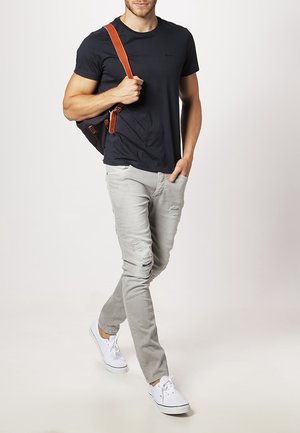 Homme marchant, portant un t-shirt noir, un jean déchiré gris clair, des baskets blanches, et portant un sac noir avec des sangles marron sur son épaule.