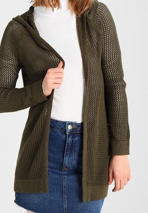 Femme portant un cardigan ajouré vert olive sur un col roulé côtelé blanc et une jupe en denim bleu, tenant l'avant du cardigan de la main.