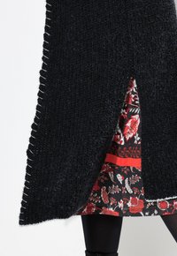 Pitkä musta neulecardigan, jossa on tekstuuri, valkoisilla ompeleilla reunat, kukkakuosisen mekon päällä, jossa on punaisia yksityiskohtia.