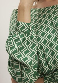 Zielona bluzka w wzory z białymi geometrycznymi wzorami. Posiada bufiaste rękawy, guzik na mankiecie oraz gładką, teksturowaną tkaninę.