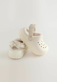 Sabots beige clair avec un intérieur doux et pelucheux. Présente des trous découpés pour la ventilation et un accent de nœud décoratif sur la sangle arrière.