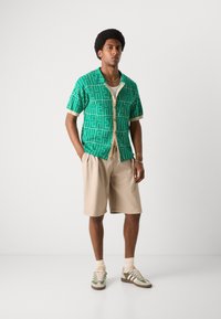 Grøn mønstret kortærmet skjorte med hvide detaljer, beige shorts og grønne/brune sneakers. Glatte teksturer, afslappet pasform, uformel stil.