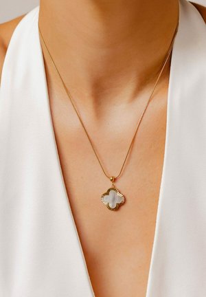 Goldene Halskette mit einem kleeblattförmigen Anhänger, der einen strukturierten Rand und einen zentralen weißen Stein aufweist, getragen über einem weißen Kleidungsstück.