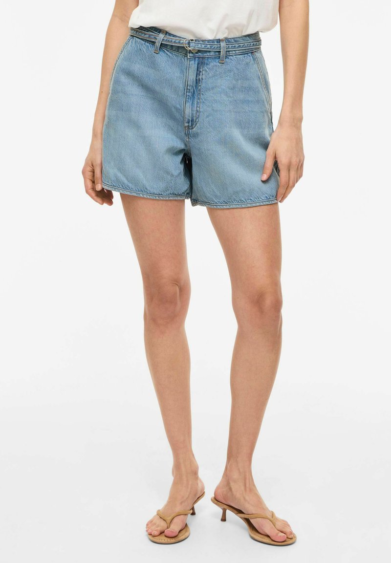 Femme portant un short en jean bleu clair avec une fine ceinture, un haut blanc et des sandales à talons beige, debout devant un fond blanc.